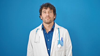 Young hispanic man doctor standing with serious expression over isolated blue background
