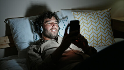 Young hispanic man watching video on smartphone lying on bed at bedroom