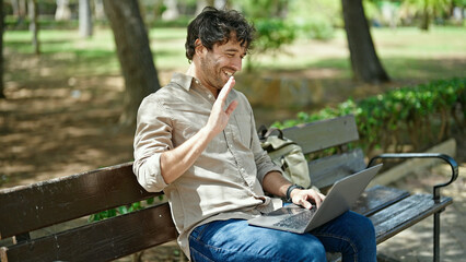 Obraz premium Young hispanic man having video call sitting on bench smiling at park