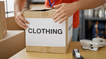 Young hispanic man volunteer packing clothes cardboard box to donate at charity center