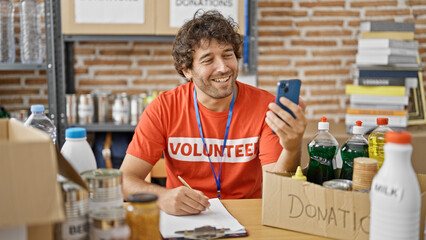 Young hispanic man volunteer writing on clipboard having video call at charity center