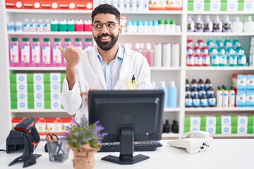 Hispanic man with beard working at pharmacy drugstore smiling with happy face looking and pointing...