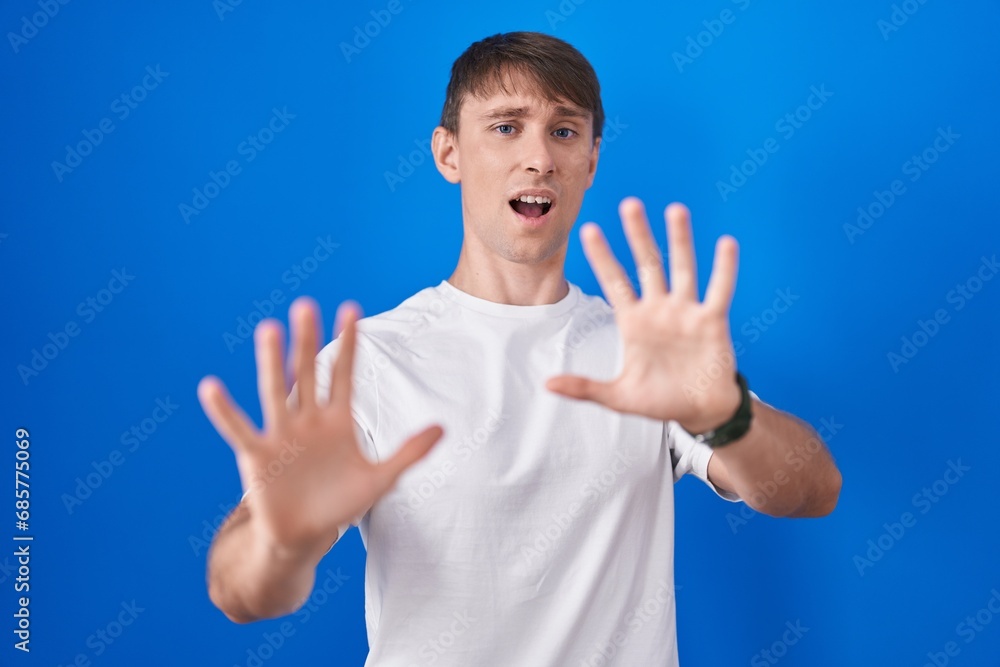 Sticker Caucasian blond man standing over blue background afraid and terrified with fear expression stop gesture with hands, shouting in shock. panic concept.