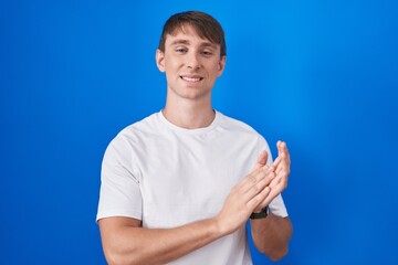 Caucasian blond man standing over blue background clapping and applauding happy and joyful, smiling proud hands together