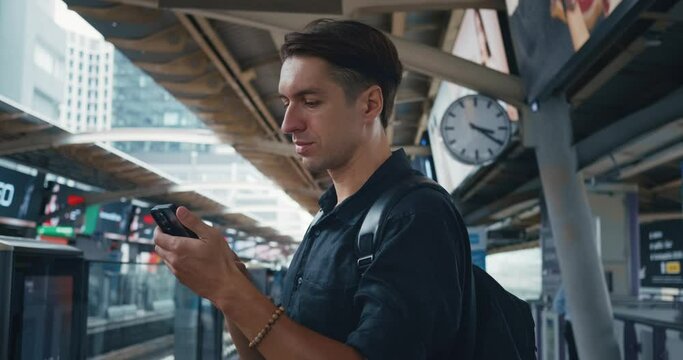 Male tourist with backpack using smartphone while waiting for metro train at metro station at modern big city. Man tourist waiting sky train at platform station of public transport. Traveling concept