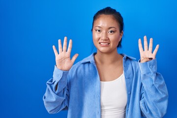 Asian young woman standing over blue background showing and pointing up with fingers number nine...