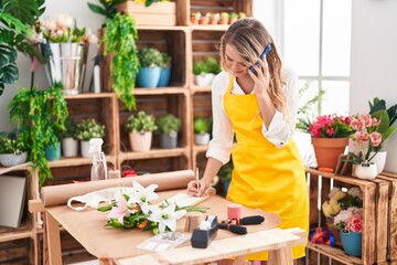 Young blonde woman florist talking on smartphone writing on notebook at florist