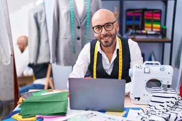 Young bald man tailor smiling confident using laptop at tailor shop