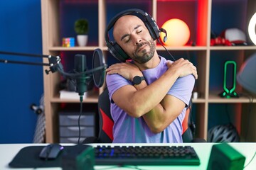 Middle age bald man playing video games wearing headphones hugging oneself happy and positive, smiling confident. self love and self care