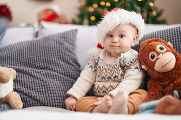 Adorable caucasian baby sitting on sofa by christmas tree with relaxed expression at home