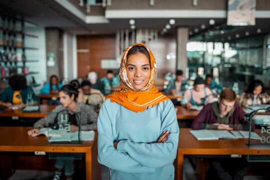 Portrait of a smiling young Arabian woman in hijab standing in library. Young female student in busy library looking at camera with confidence