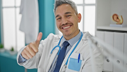 Young hispanic man doctor doing thumb up sitting on table at clinic