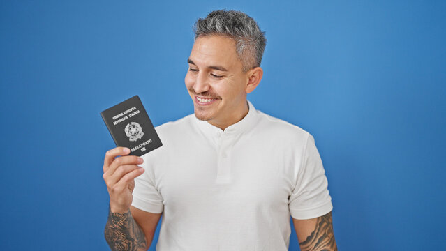 Young hispanic man smiling confident holding italian passport over isolated blue background