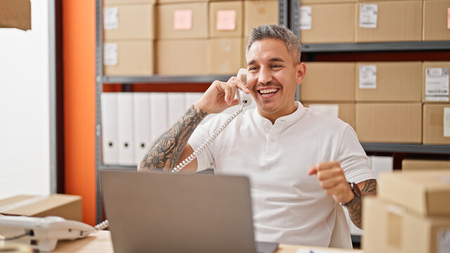 Young hispanic man ecommerce business worker using laptop talking on telephone celebrating at office