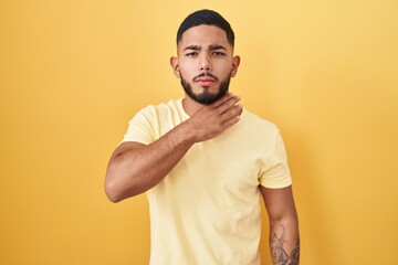 Young hispanic man standing over yellow background cutting throat with hand as knife, threaten aggression with furious violence