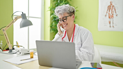 Young woman doctor using laptop talking on telephone at clinic