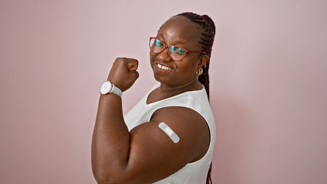 Confident african american woman with braids makes strong gesture, band aid on arm, expressing joy over isolated pink background amidst vaccination