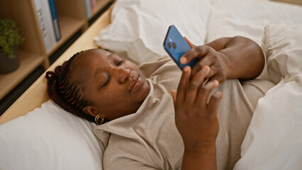 Relaxed african american woman comfortably utilizing her smartphone while lying in bed at home, surrounded by cosy bedroom interior