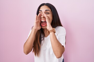 Young arab woman standing over pink background shouting angry out loud with hands over mouth