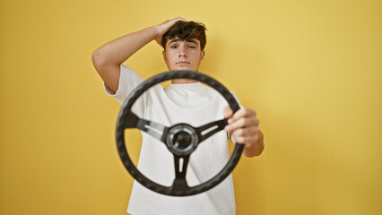Young hispanic teenager, a serious yet upset novice driver, struggling with steering wheel against isolated yellow wall background