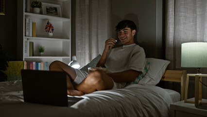 Enthralled young hispanic teenager, comfortably sitting on his bed, engrossed in watching a movie on his laptop, radiating a positive vibe in his cozy bedroom at night, making every pixel count.