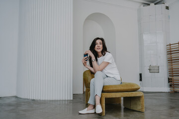 Relaxed brunette woman in casual clothe sitting at minimalistic interior holds phone looks aside with pensive face. Lonely young woman at home waiting for message from boyfriend.