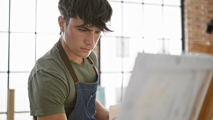 Serious young hispanic teenager artist engrossed in drawing portrait at indoor art studio, standing...