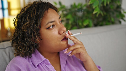Young beautiful latin woman sitting on sofa smoking cigarette at home