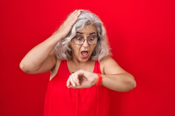 Middle age woman with grey hair standing over red background looking at the watch time worried,...