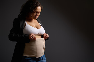 Authentic woman, young mother putting on elastic bandage after c-section, isolated on gray studio...