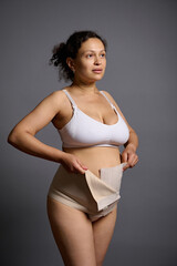 Multi ethnic woman, young mother putting on a bandage after c-section, looking away, isolated on...