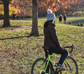 woman holding bicycle looking away from camera in park (dressed i winter hat, brown puffy jacket, dark long hair) sun flares, haze, autumn and winter light (sunset, glow) nature, cycling, gravel bike
