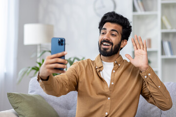 Happy young man hold smartphone say hello talking by online video call with family or friends on couch at home Handsome male greeting to someone looking at cellphone screen indoors High quality photo