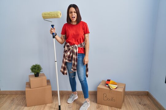Young hispanic woman painting home walls with paint roller angry and mad screaming frustrated and furious, shouting with anger. rage and aggressive concept.