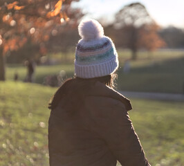 woman with long dark hair shot from behind looking into field in a park (no face, winter hat, warm autumn jacket) single lonely isolation look sunset sun flare