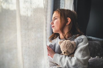 a pensive cute girl sits and looks out the window, hugging a teddy bear. - Powered by Adobe