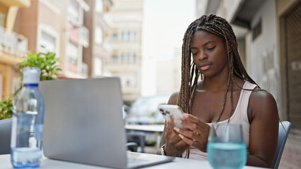 African american woman using laptop and smartphone sitting on table at coffee shop terrace