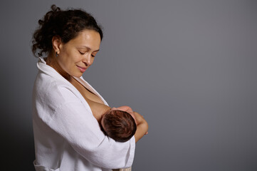 Smiling beautiful woman enjoys breastfeeding her baby, isolated over gray studio background.