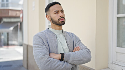 Young hispanic man standing with serious expression and crossed arms at street