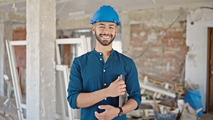 Young hispanic man architect wearing hardhat smiling holding laptop at construction site