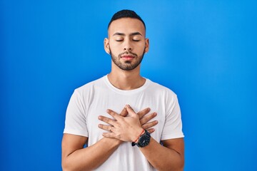 Young hispanic man standing over blue background smiling with hands on chest with closed eyes and grateful gesture on face. health concept.