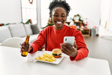 Fototapeta na wymiar African american woman eating hamburger using smartphone sitting on table by christmas tree at home