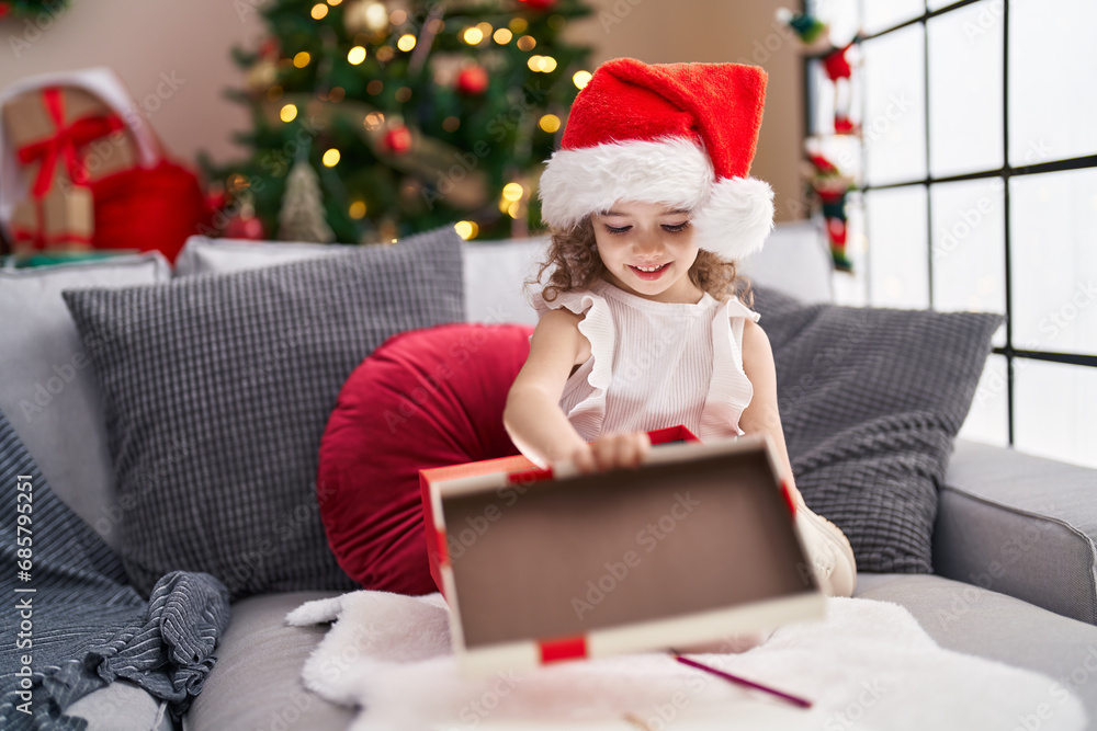 Poster adorable caucasian girl unpacking gift sitting on sofa by christmas tree at home