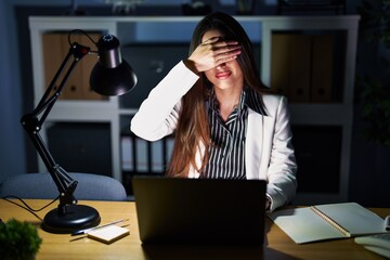 Young brunette woman working at the office at night with laptop covering eyes with hand, looking serious and sad. sightless, hiding and rejection concept