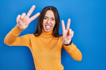 Young brunette woman standing over blue background smiling with tongue out showing fingers of both hands doing victory sign. number two.