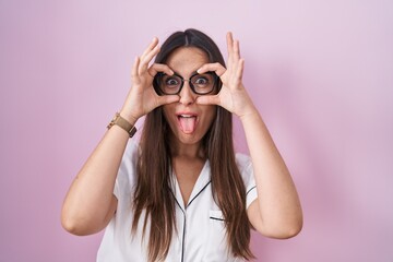 Young brunette woman wearing glasses standing over pink background doing ok gesture like binoculars sticking tongue out, eyes looking through fingers. crazy expression.
