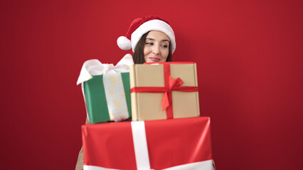 Beautiful hispanic woman wearing christmas hat holding gifts over isolated red background