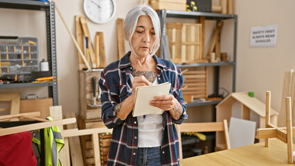 Serious mature grey-haired female carpenter taking detailed notes in her lively woodworking studio