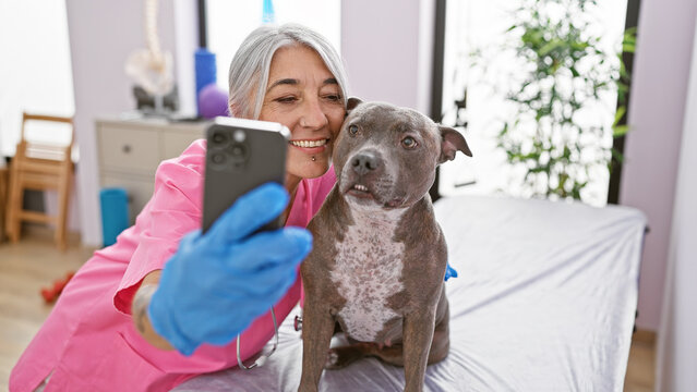 Smiling middle-aged veterinarian adorably taking selfie with a senior dog at vet clinic, immersed in the world of pet health care