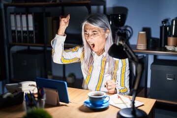 Middle age woman with grey hair working at the office at night angry and mad raising fist...